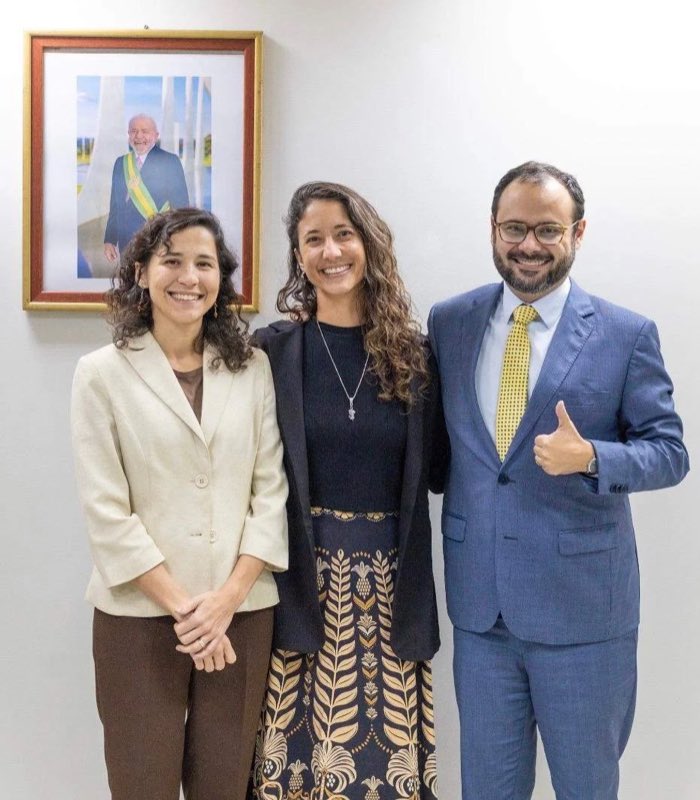 Natália Rinco entre duas pessoas em um gabinete do governo federal, com o retrato oficial do presidente da República ao fundo.