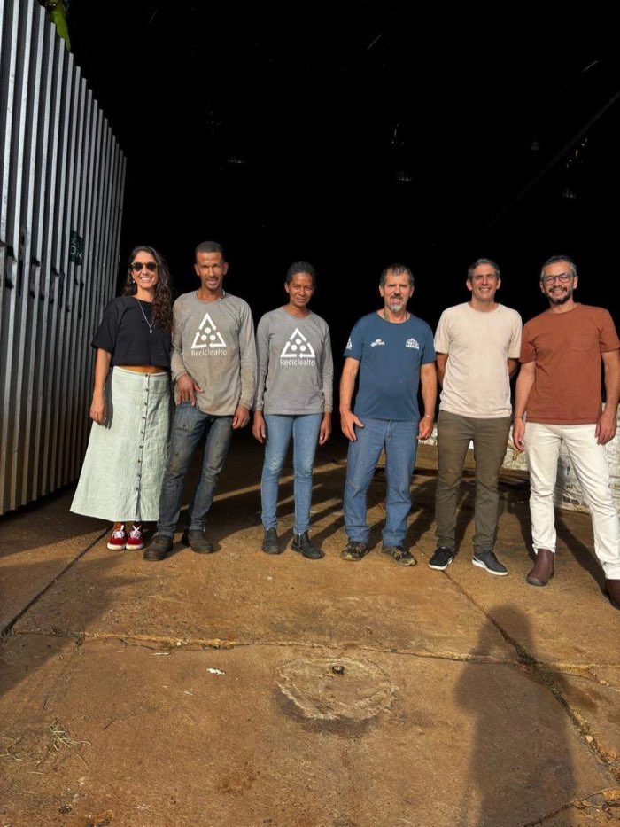 Natália Rinco ao lado de trabalhadores de camiseta da Reciclealto, dentro do galpão de triagem, com fardos de material reciclável ao fundo.